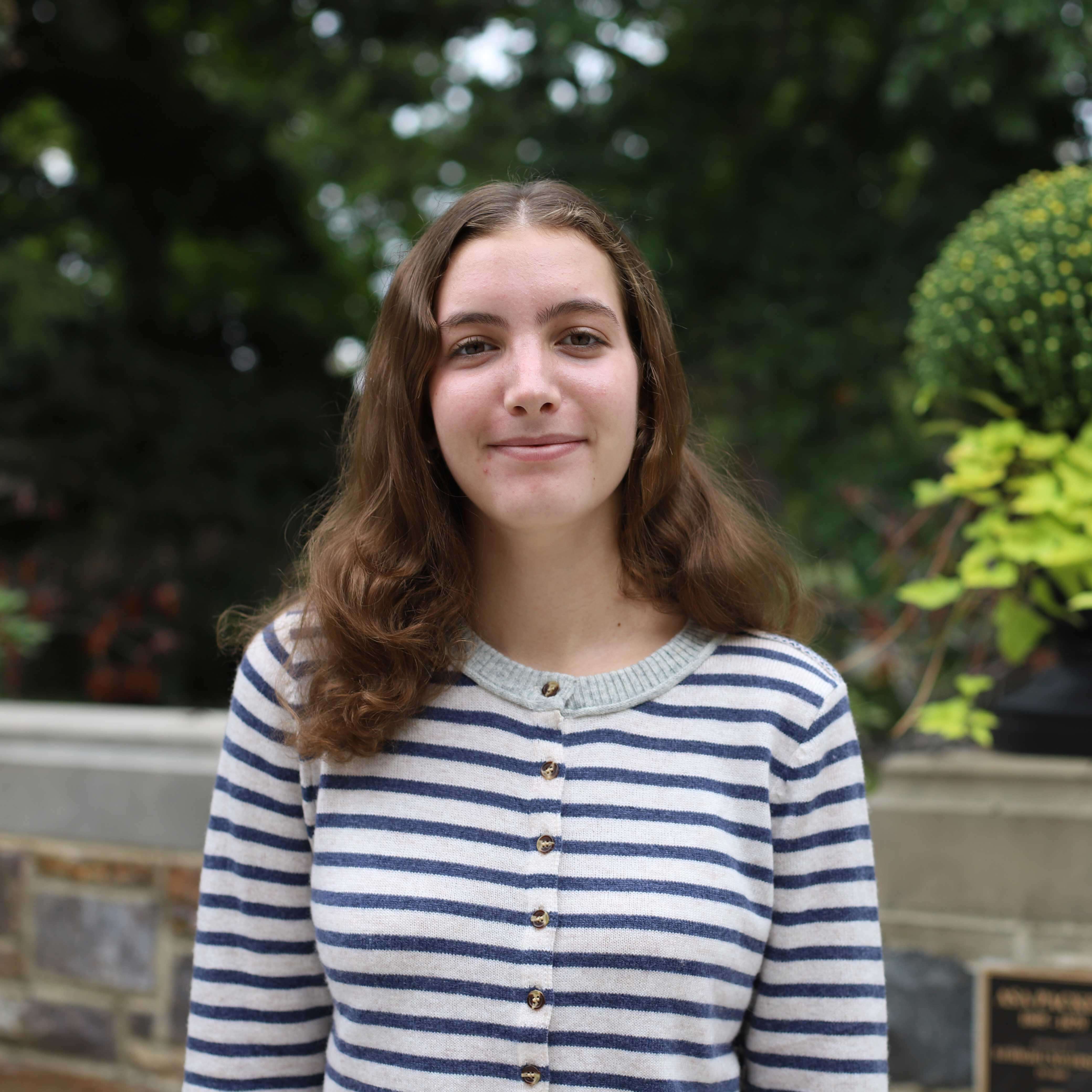 Rebecca wears a blue and white striped sweater. Trees and greenery are visible behind her.
