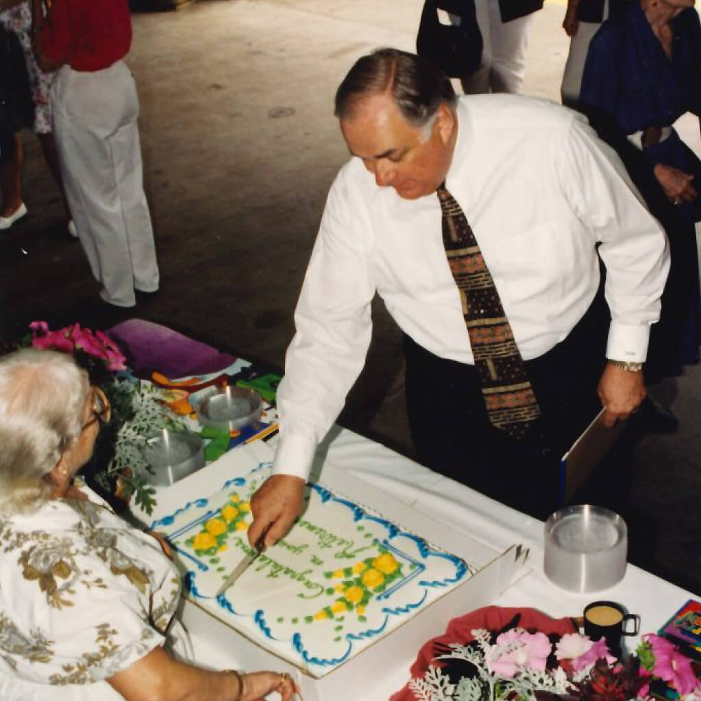 David Van Horn, wearing a white dress shirt and striped neck tie, leans over a table with a cake, dessert plates, and flowers and prepares to cut into the cake.