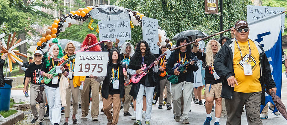 A group of alumni parade with long wigs and signs that read "Class of 1975" and "Still Rockin' and Rollin”