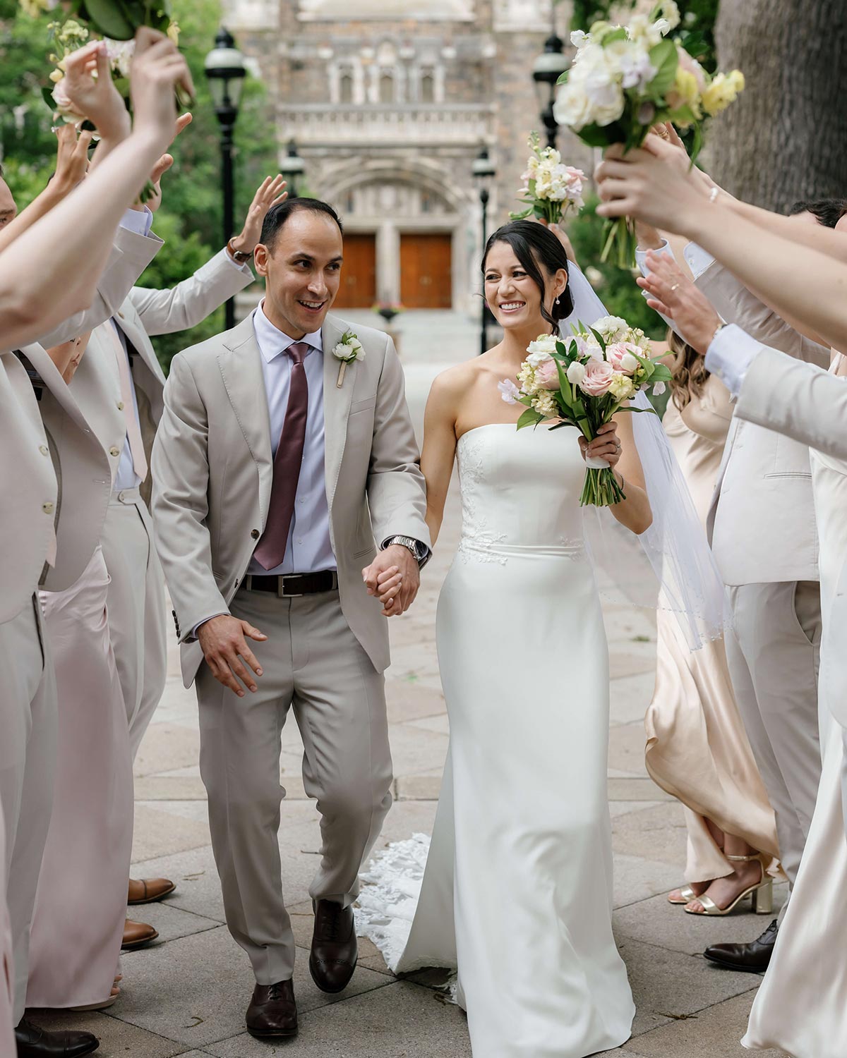 Cassidy and Ricardo walk by their cheering friends after their wedding; she wears a strapless gown and he wears a tan suit.