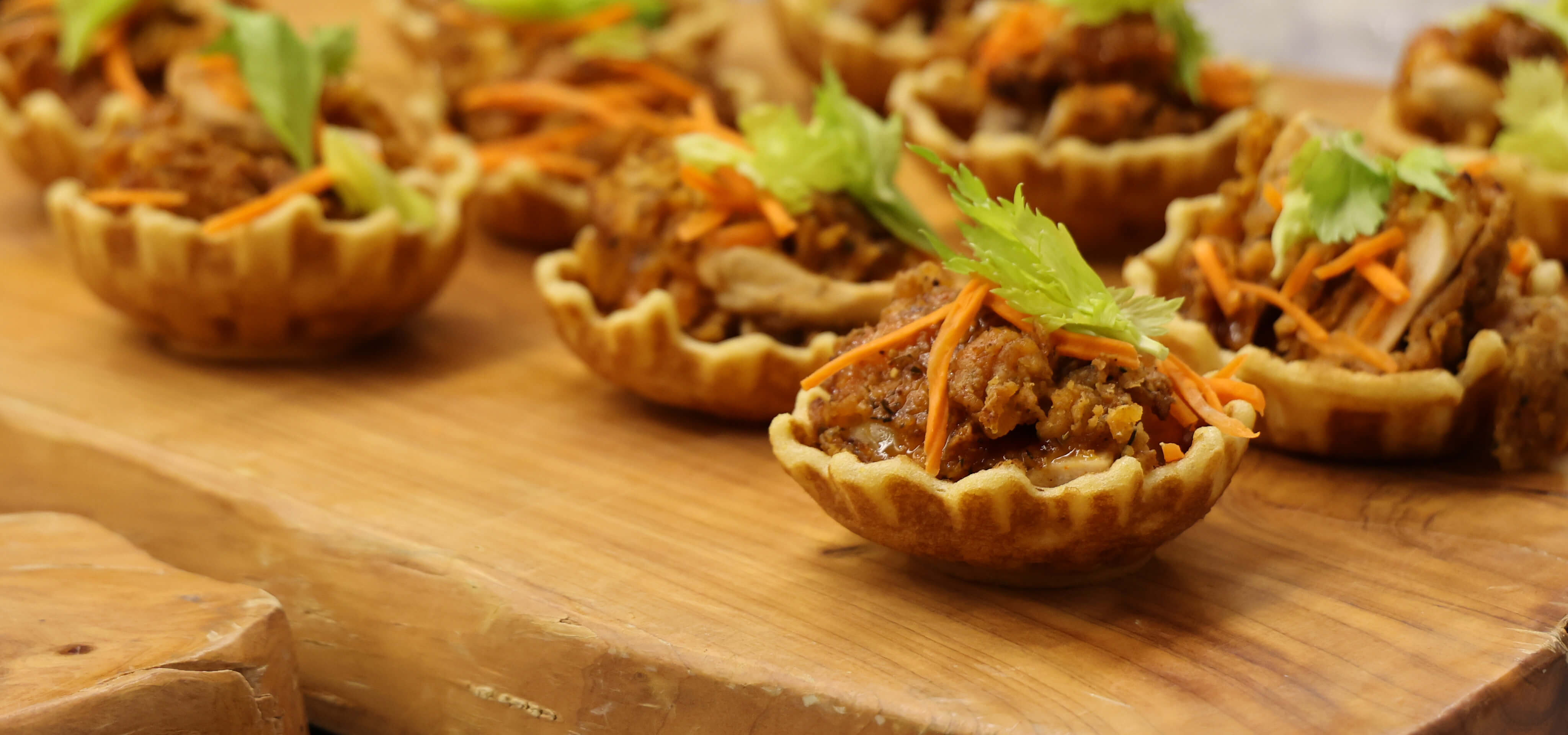 A buffet of small waffle bowls filled with chicken and topped with carrot and green garnish sit atop a rustic wooden cutting board.