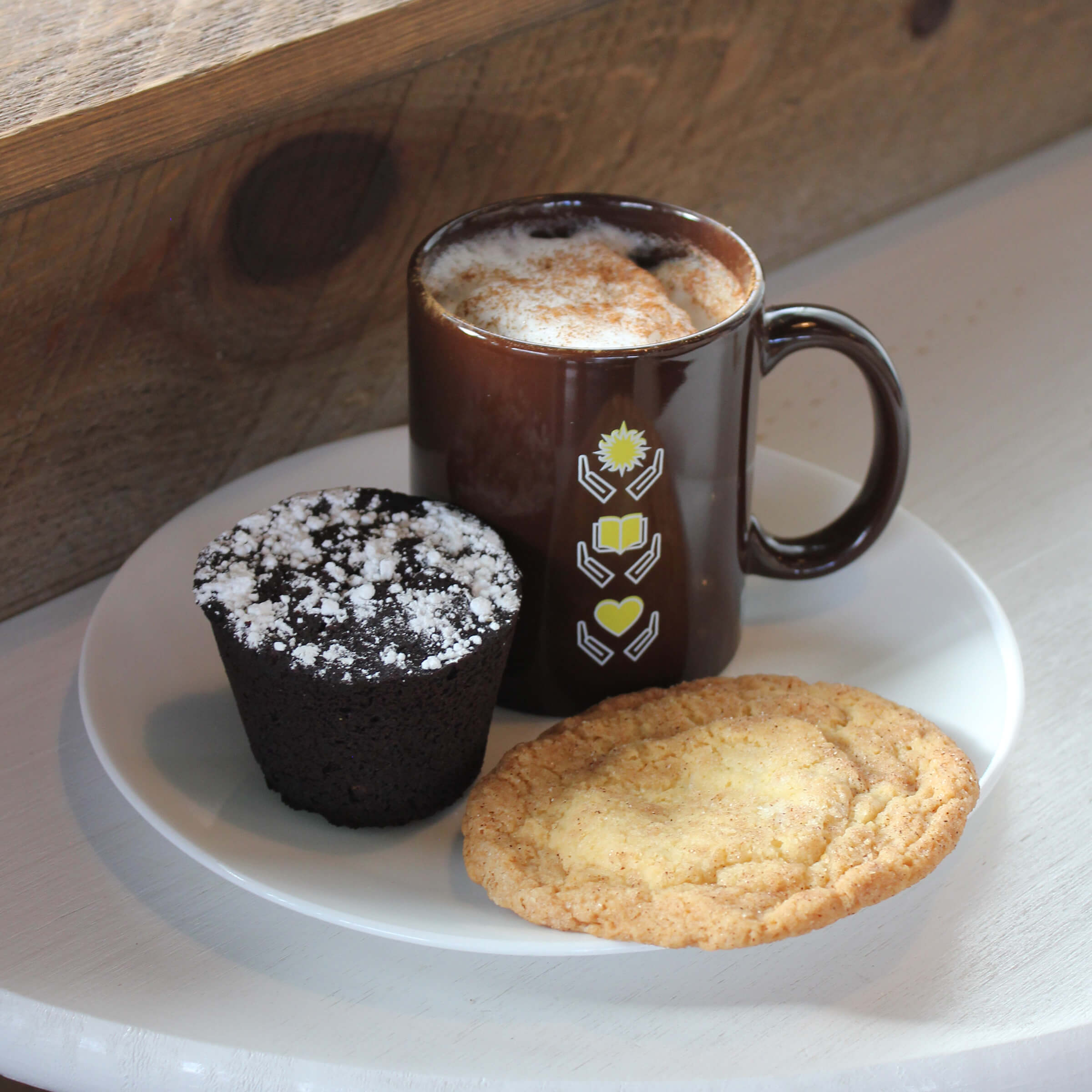 A latte in a brown mug topped with froth and cinnamon flanked by a snickerdoodle cookie and tall brownie dusted with sugar.