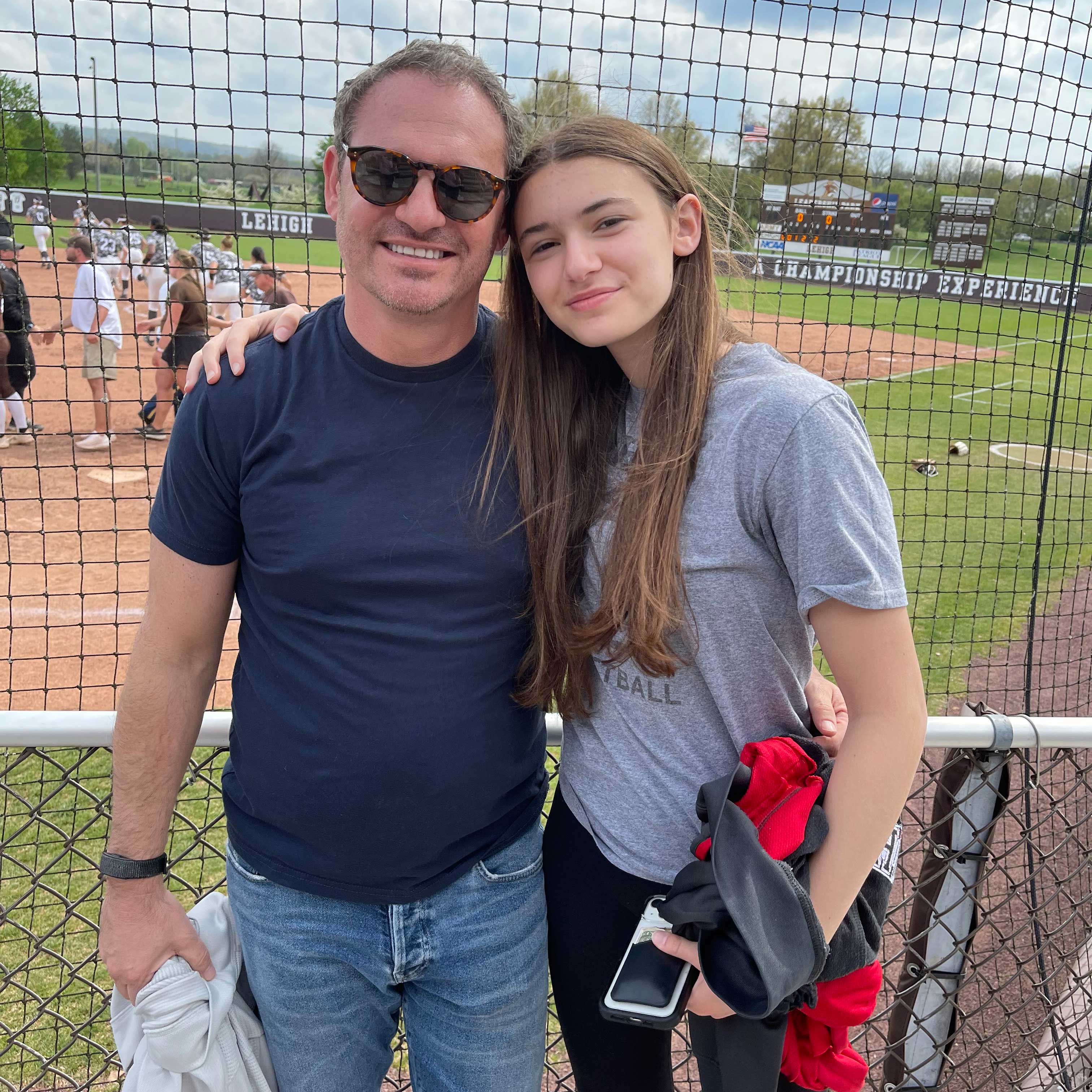 Rob stands with his daughter, Emme, on a bright, sunny day. Lehigh's baseball field is visible behind them.