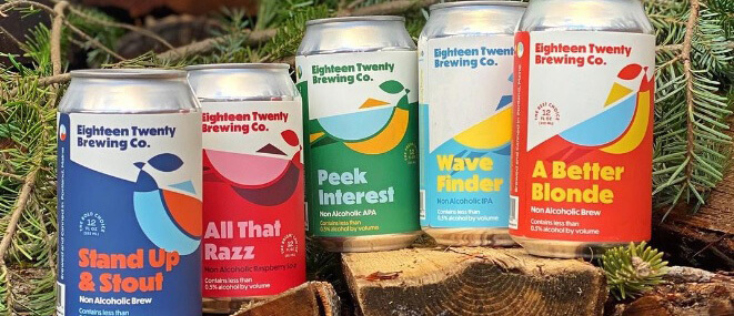 Five colorful cans of non alcoholic beverages sit atop logs of wood surrounded by pine branches.