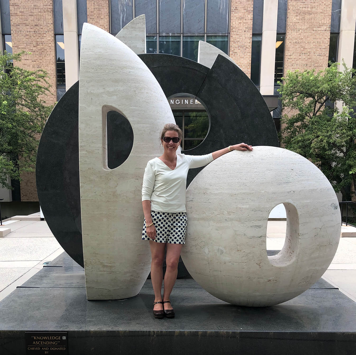 Robin Lapoint wearing sunglasses poses with “Knowledge Ascending” sculpture with one arm resting on the sculpture.