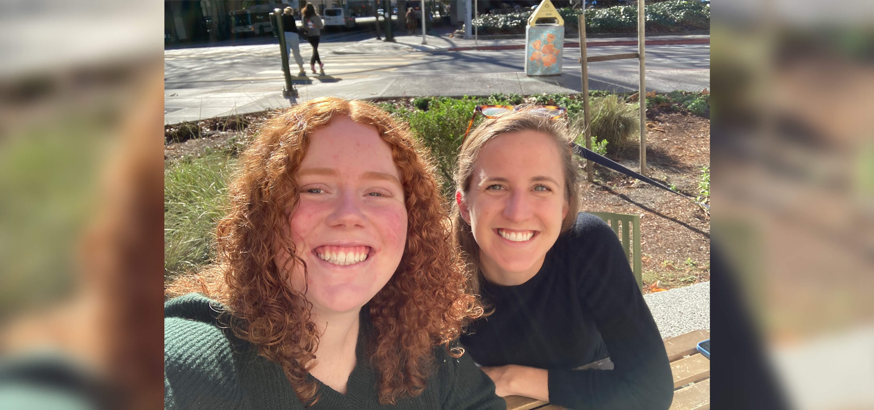 Two women pose for a selfie while sitting outside on a park bench and smiling.
