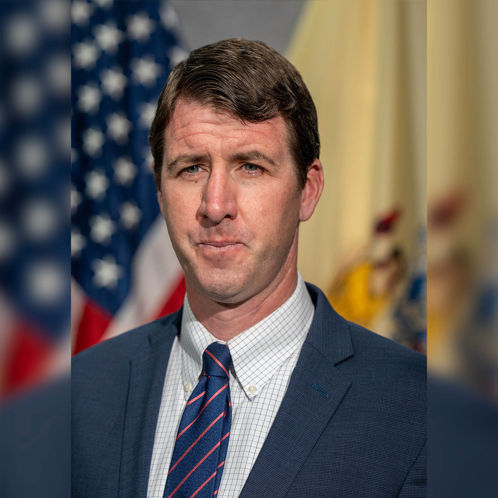 A man with brown hair wears a blue suit jacket and blue tie with red stripes while posing in front of American flag and looking off camera.