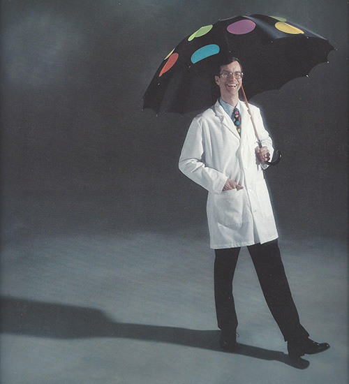 Ron wears a white lab coat and holds a polka dot umbrella