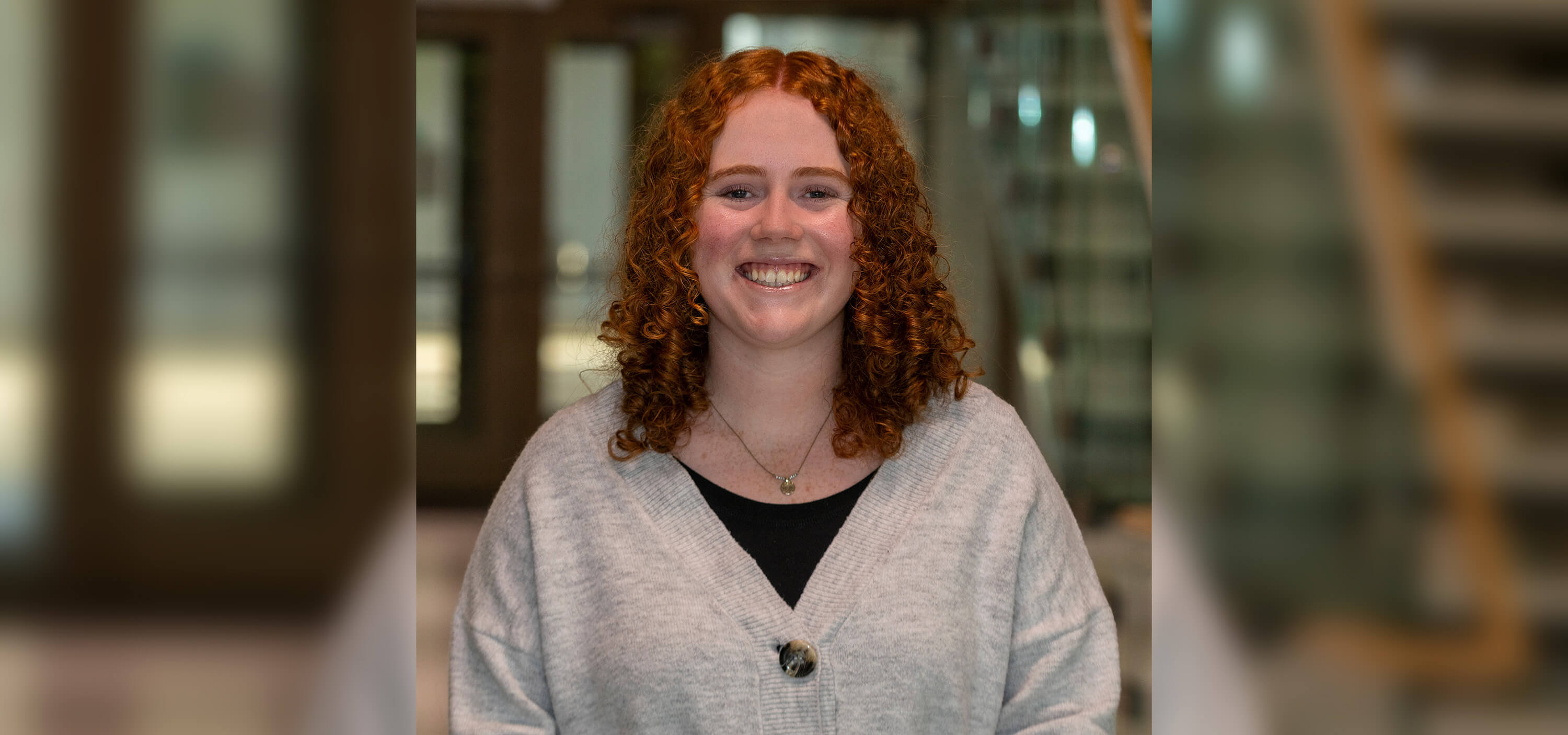 Female student with curly red hair wearing a grey cardigan over a black top smiles for a headshot.