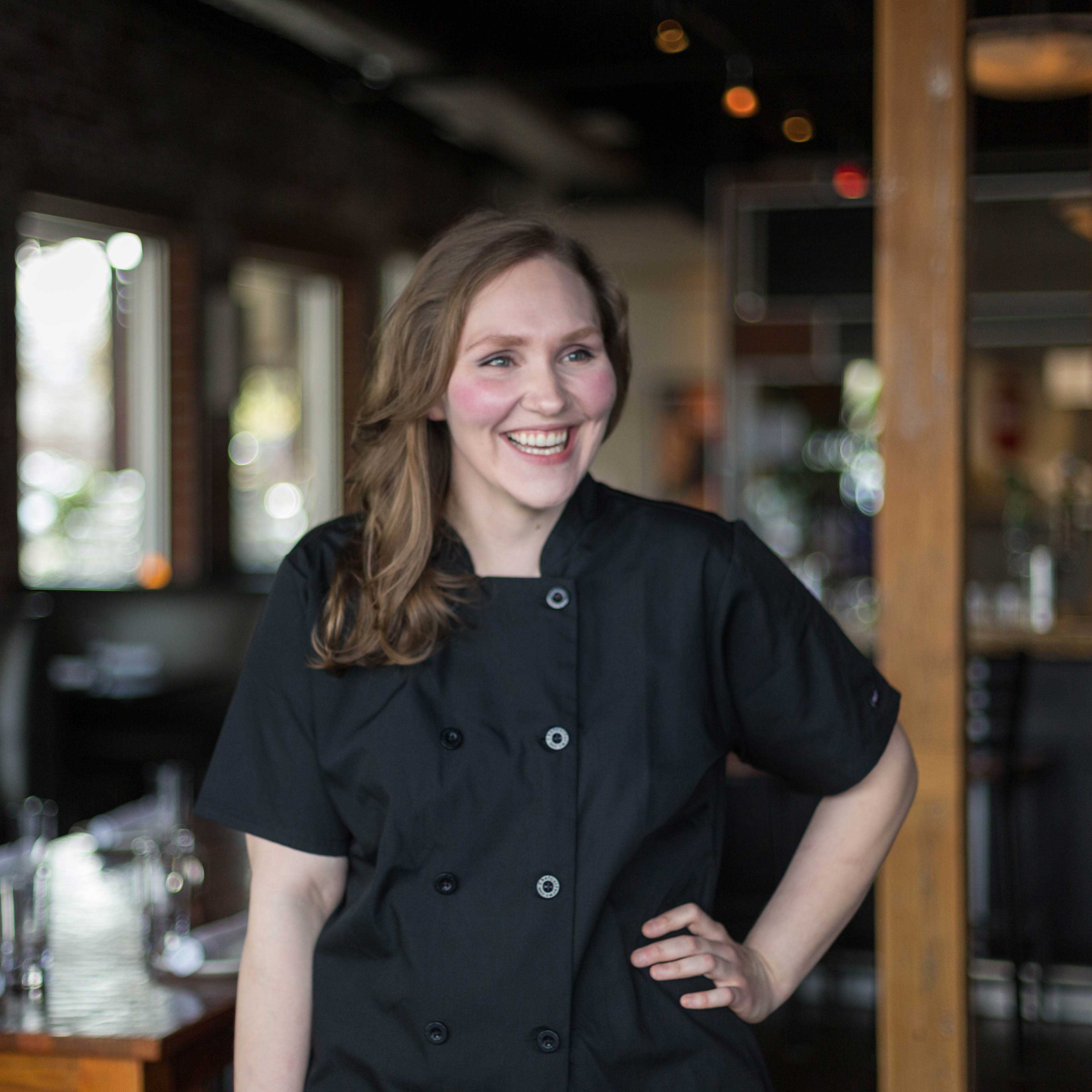 Martina wears a black chef uniform and smiles with her right hand on her hip. She stands inside a restaurant. 