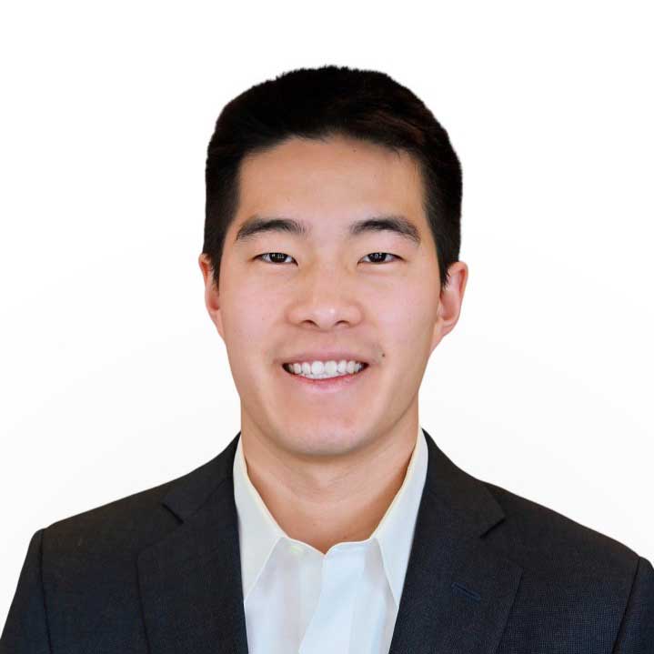 Ryan wears a black suit jacket with a white button up underneath. He stands in front of a white background and smiles.
