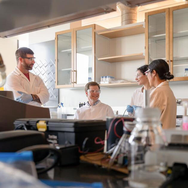 Four scientists wearing safety goggles stand together talking in a lab, with bottles and beakers shown in the foreground.