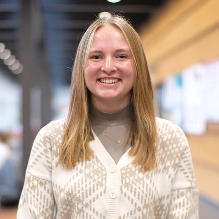 Student Olivia Meyer poses in an indoor space on campus, smiling at the camera and wearing a white sweater over a tan mock turtleneck.