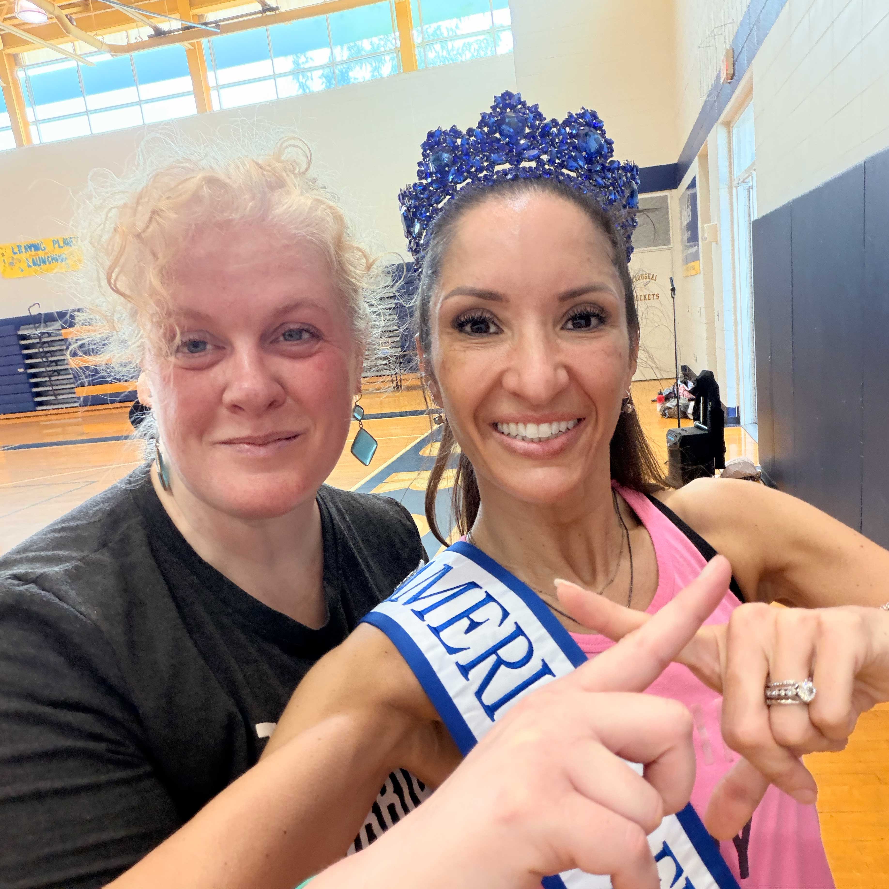 Sabrina and Kate pose in Broughal's gym. They both wear athletic clothing; Kate wears a blue crown and sash.