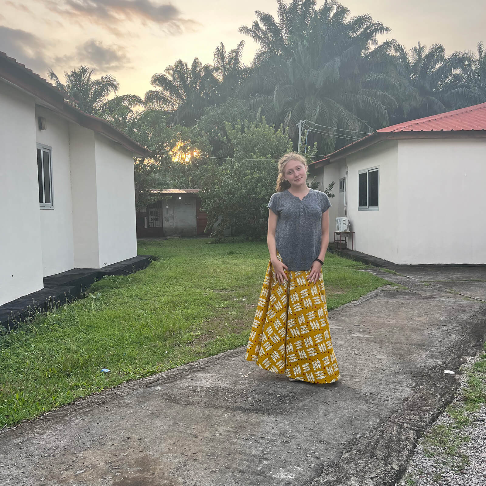 Standing on a village path with tall palm trees and white houses in the background, Samantha Sandhaus stands wearing a gray shirt and long yellow skirt.