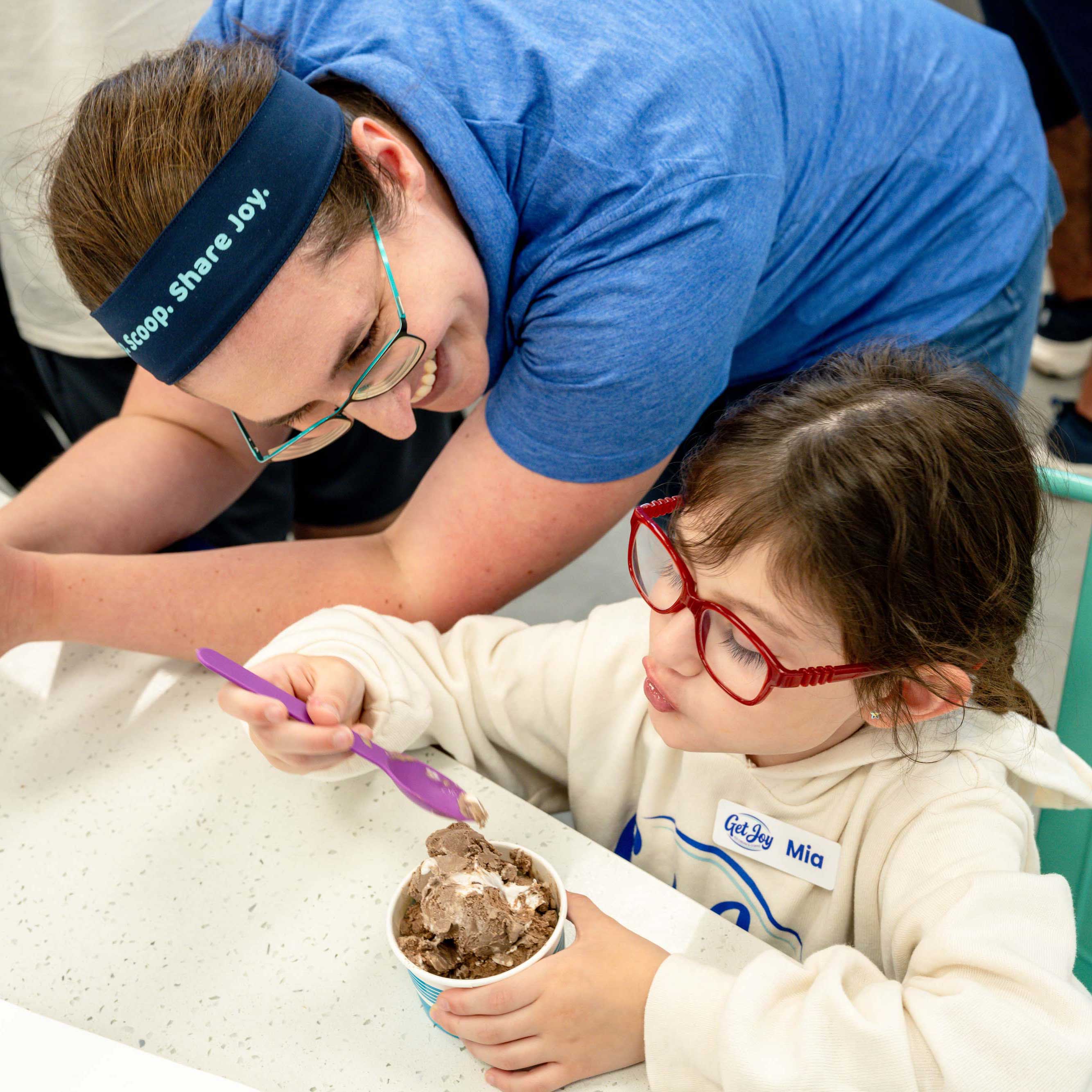 Sara leans across a table and smiles at a child eating ice cream at her shop's opening. 