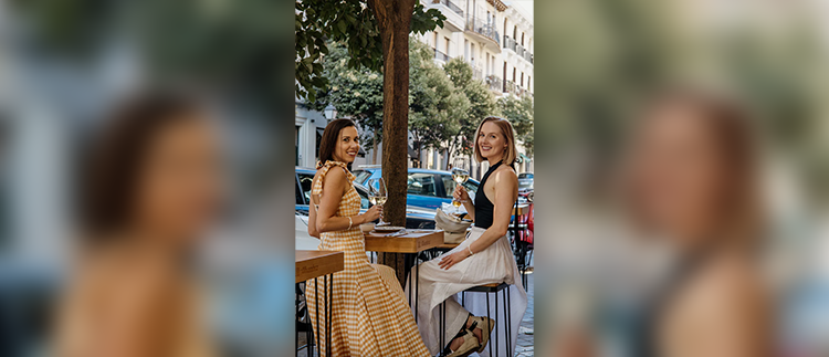 Two women sit outside at a high top table holding glasses of wine.