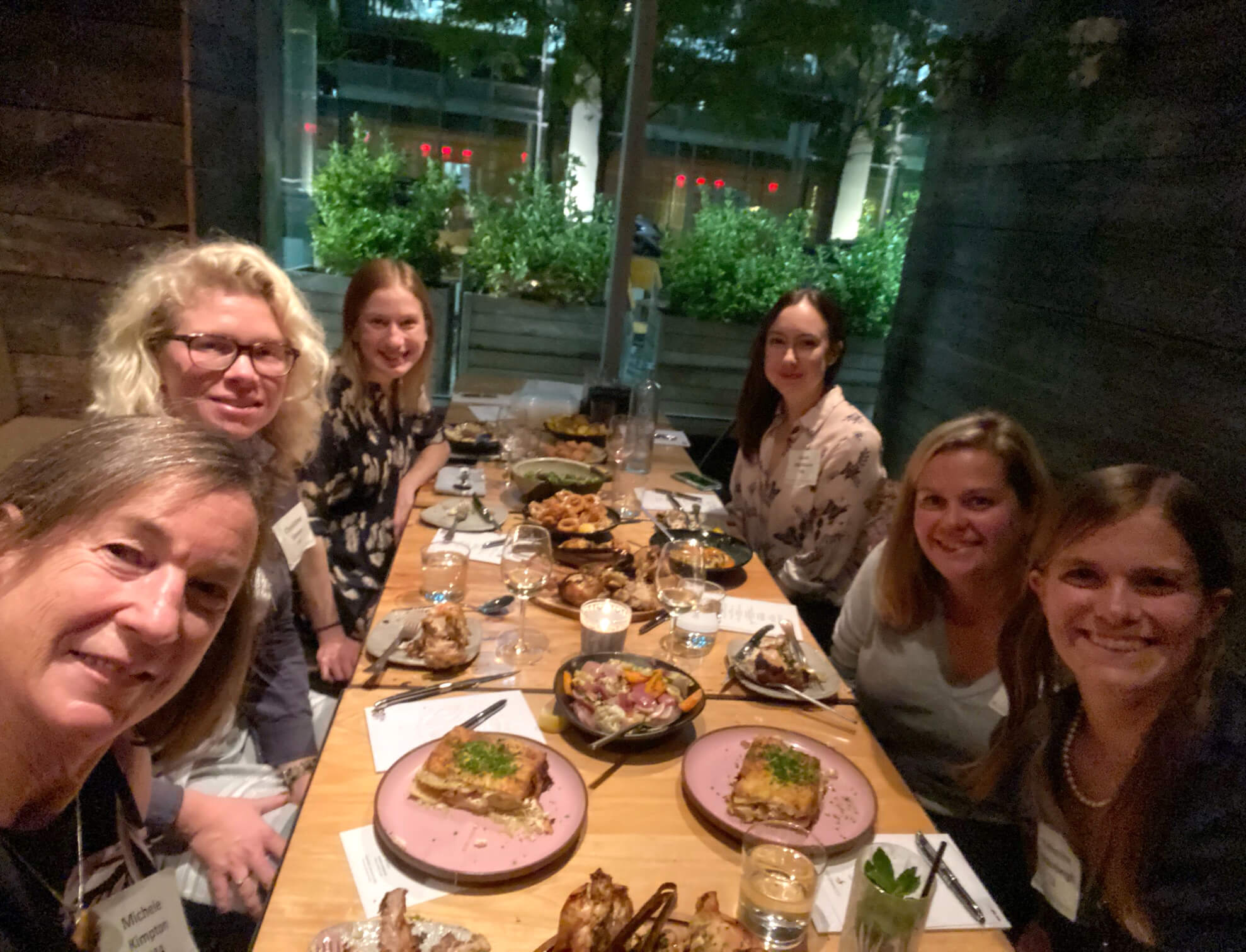 Six women sit at a table in a dimly lit restaurant while posing for a selfie.