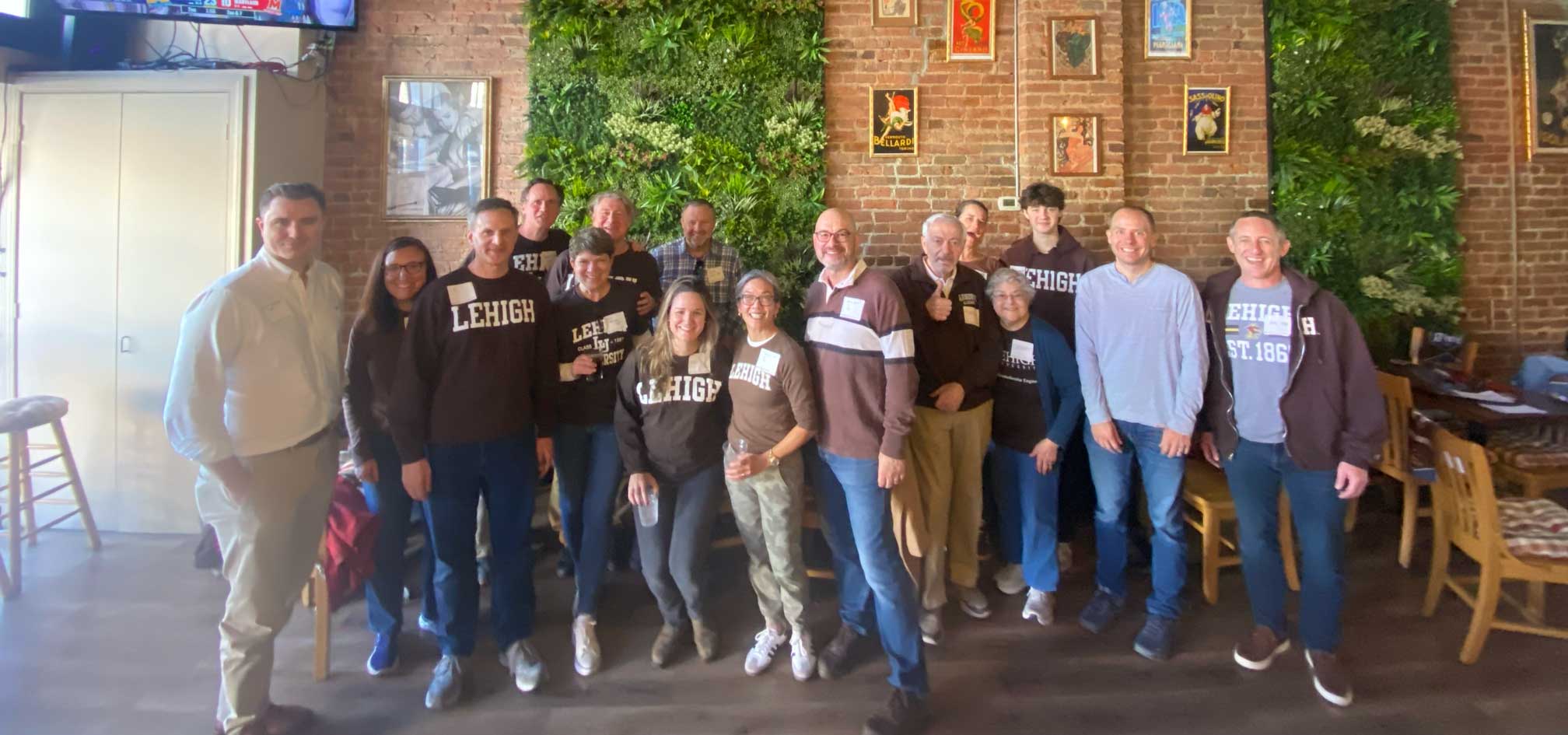 Sarah stands with a group of alumni wearing Lehigh gear while the football game plays on a television behind them.