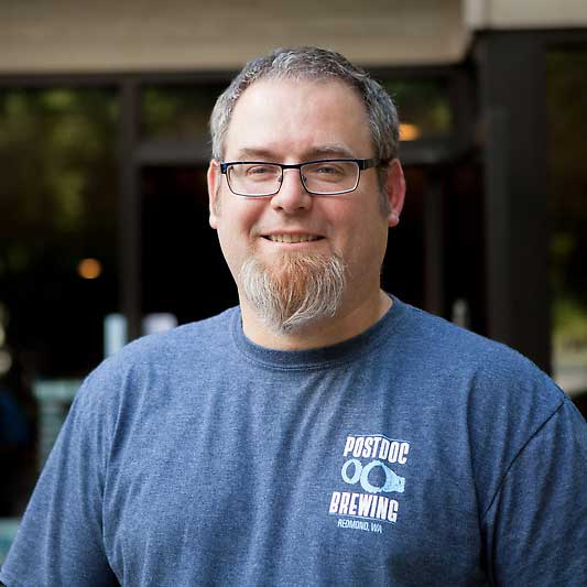 Tom wears glasses and a blue t-shirt with Postdoc Brewing's label in the upper right corner. He stands outside his brewery.