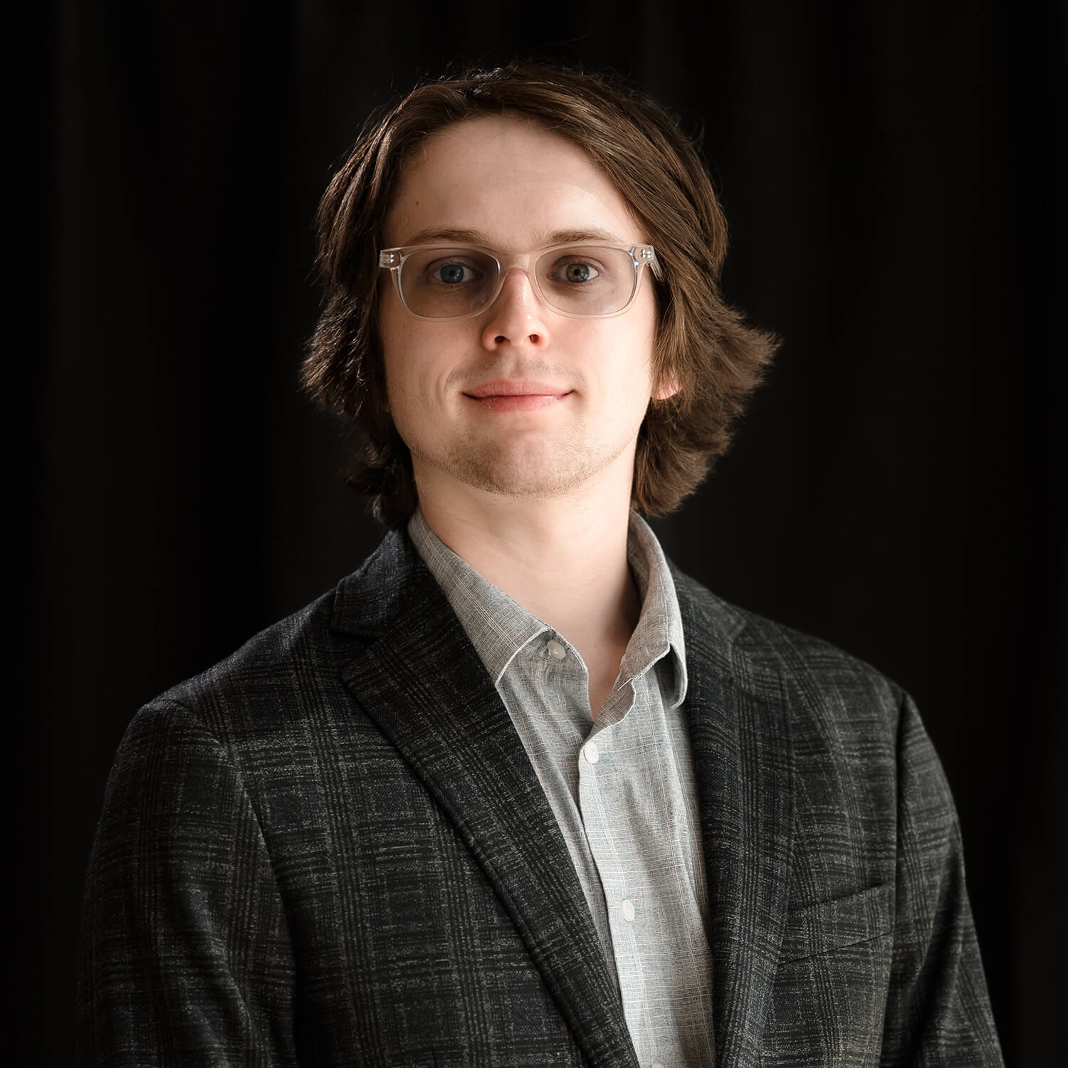On a black backdrop, Sean Henry poses with a closed-mouth smile wearing glasses, a dark gray blazer, and a lighter gray collared shirt.