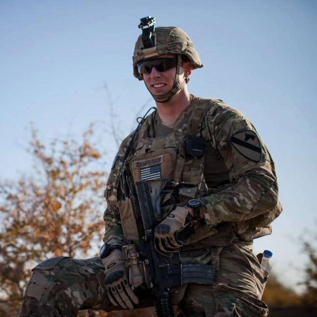 Sean wears his military fatigues, helmet, and black sunglasses while standing outside.