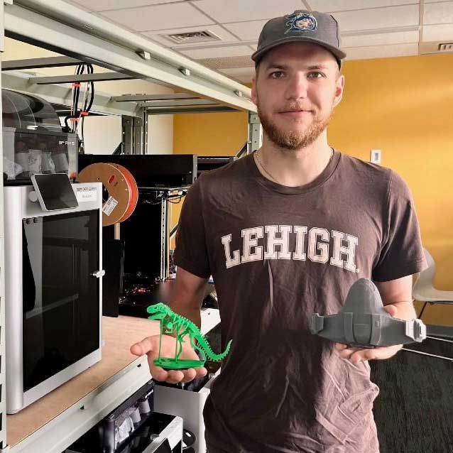 Simeon stands in the Design labs wearing Lehigh merch. He holds a 3D printed T-Rex in one hand, and a spaceship in the other.
