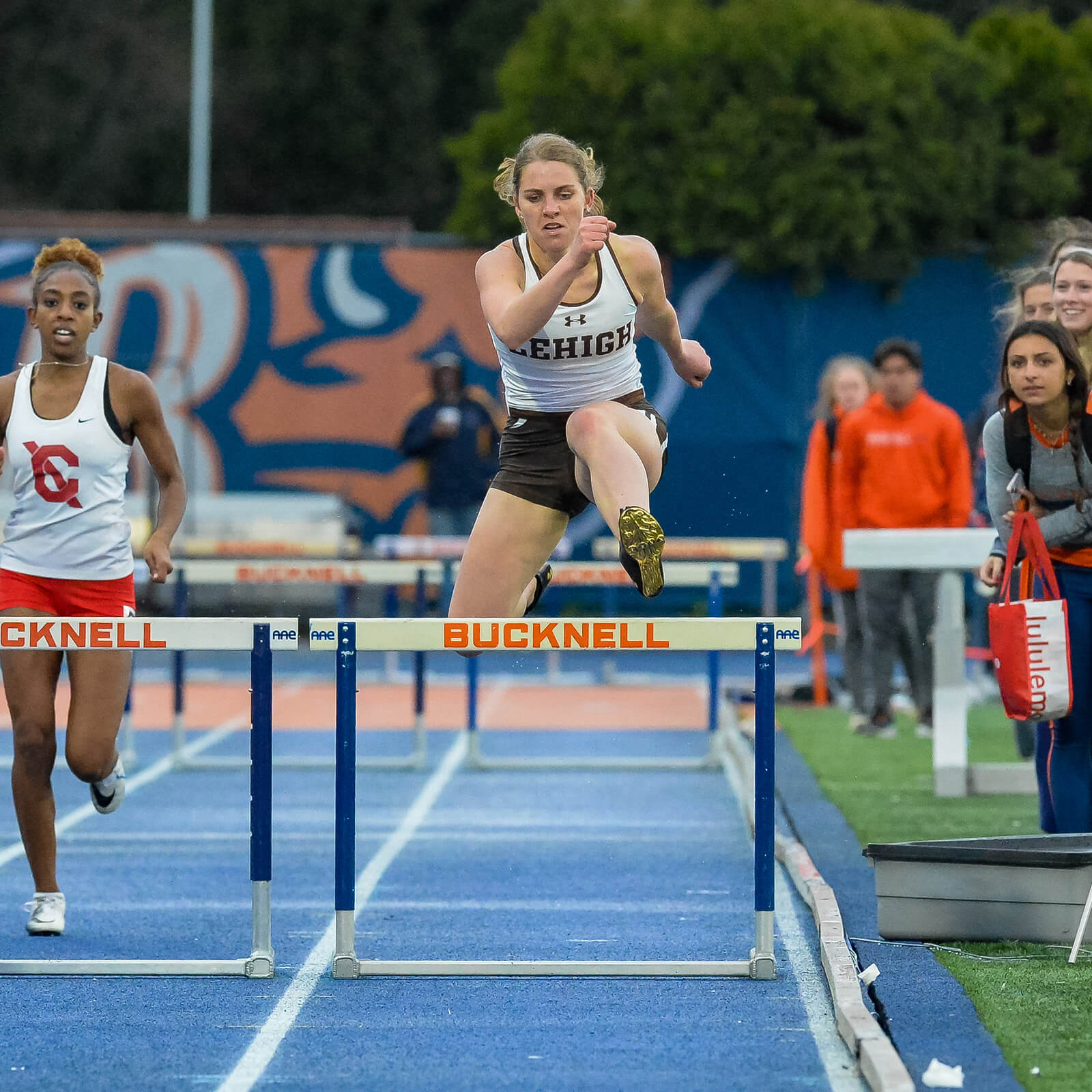 Kaja Skerlj leaps over a hurdle while racing alongside competitors on a track, wearing a white Lehigh track uniform.