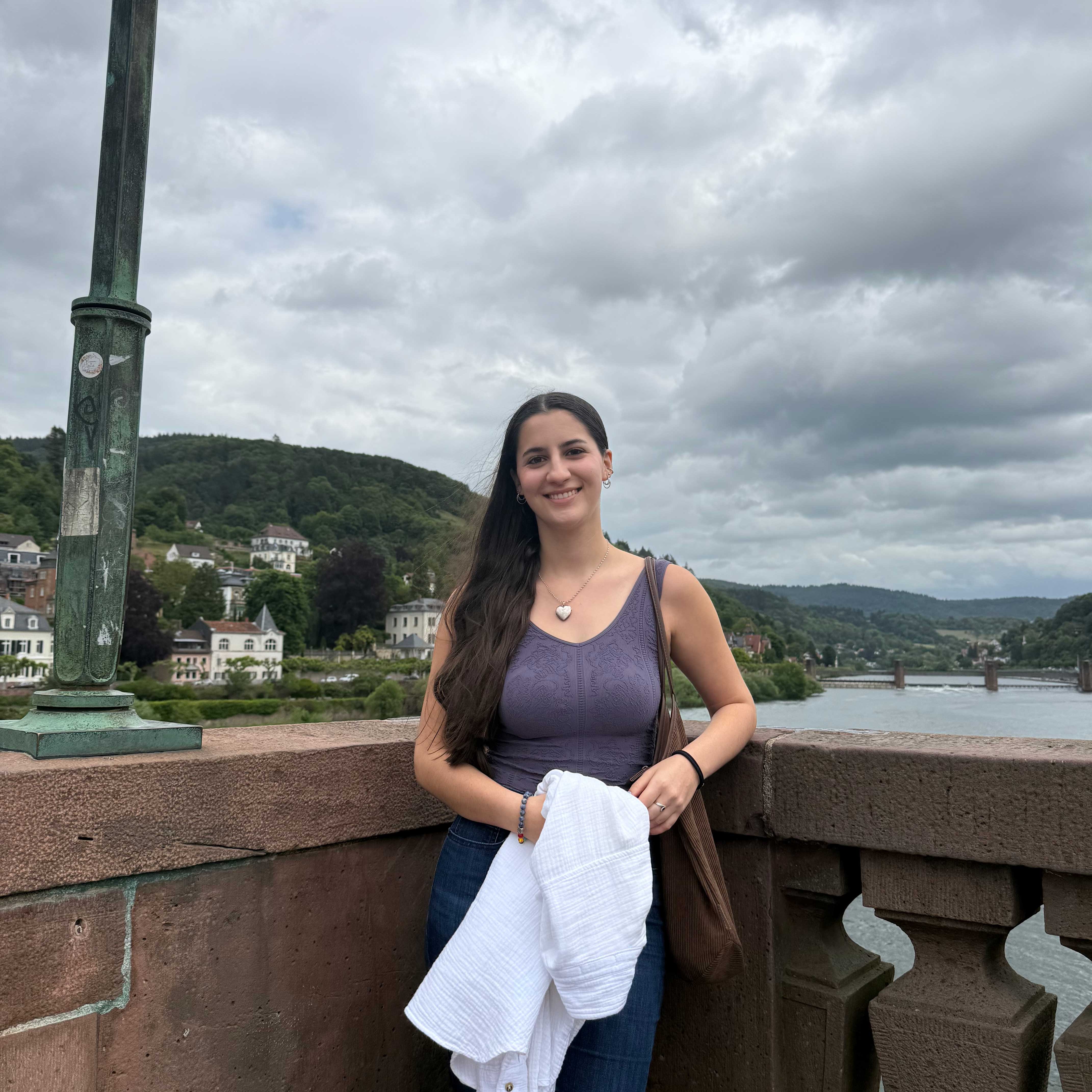 Sofia in Germany with the city visible behind her. She wears a purple top, holds a white sweater, and smiles.
