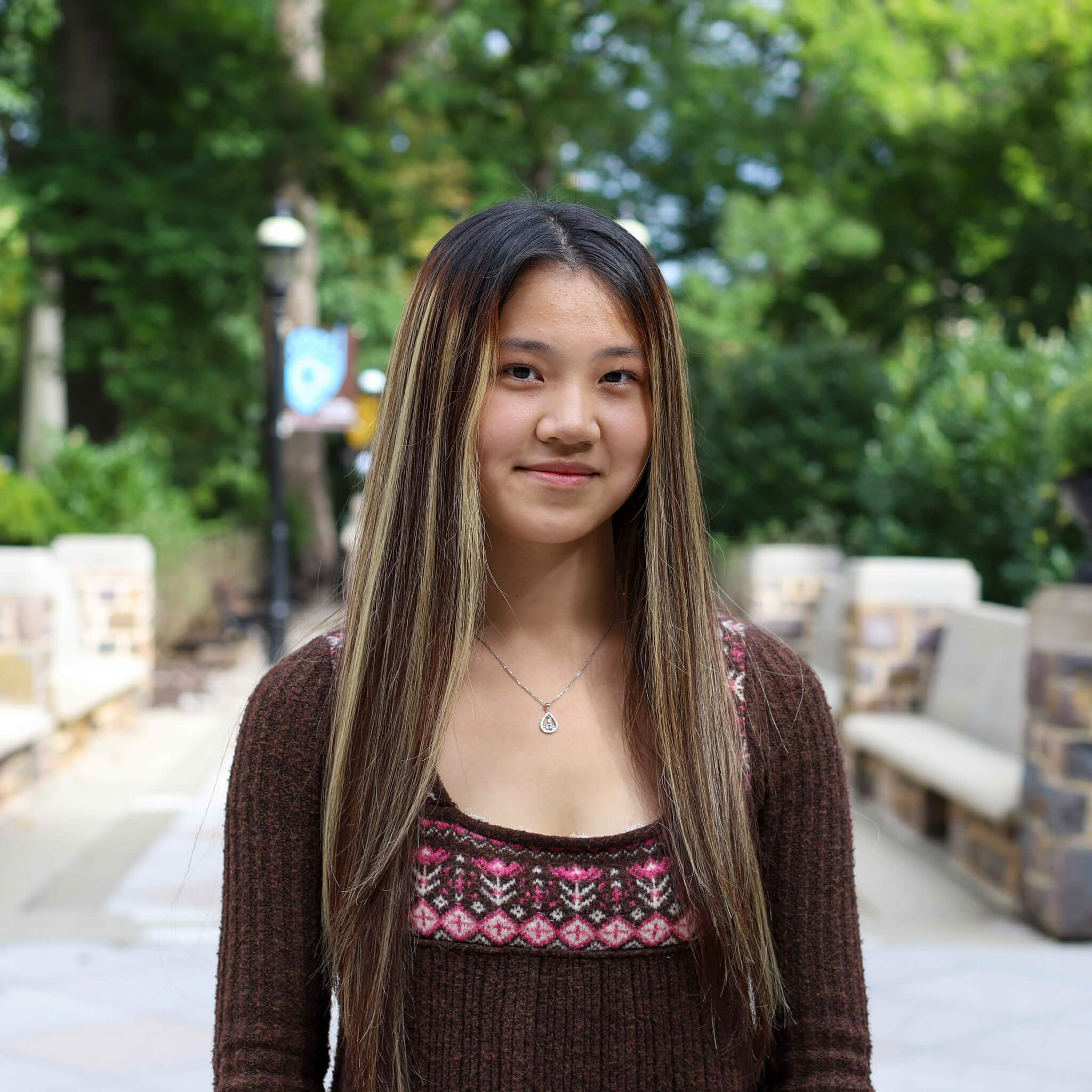 Sophia wears a brown and pink sweater and a silver necklace. Campus is visible behind her.