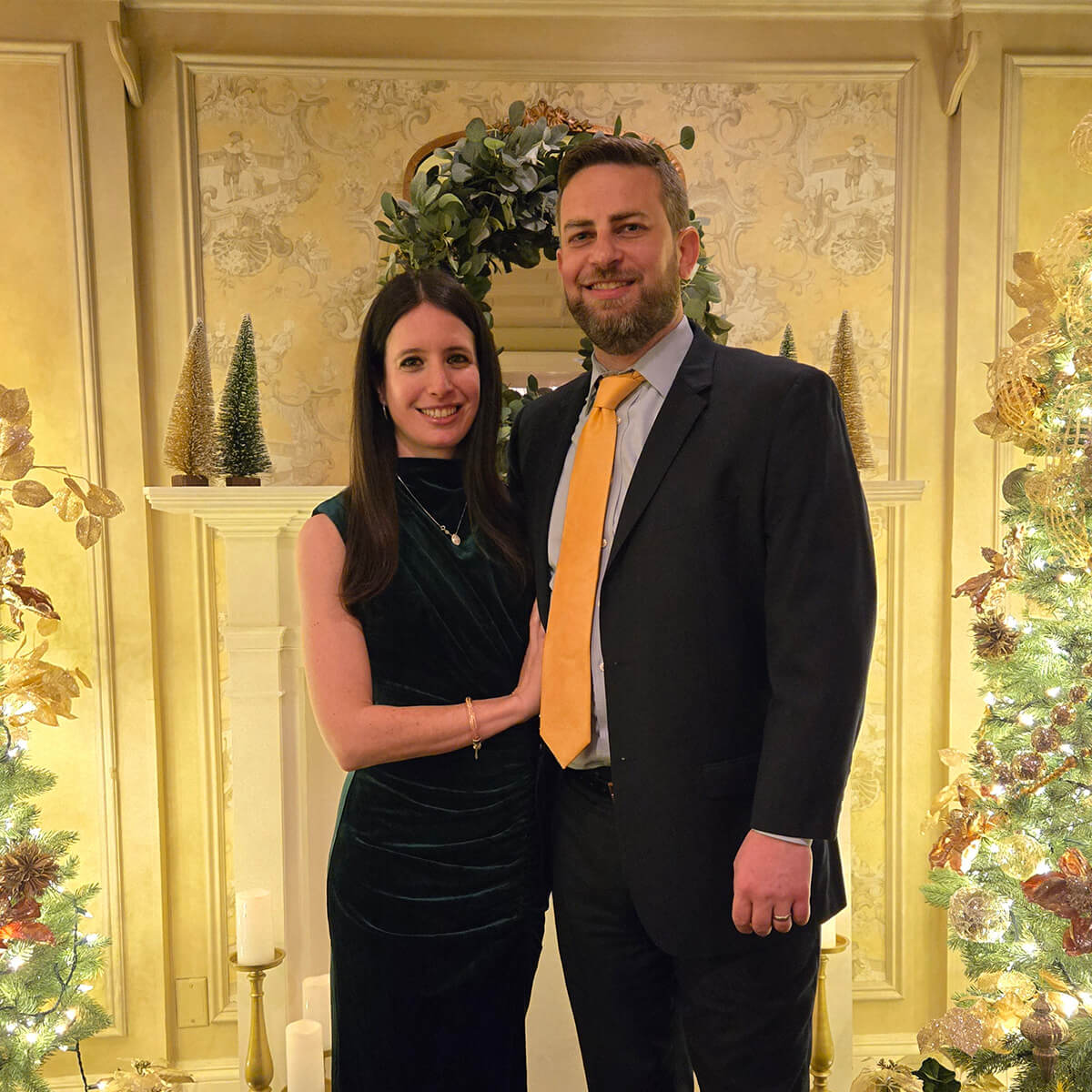 A woman and man pose in front of a holiday themed fireplace dressed in semiformal wear for an event.