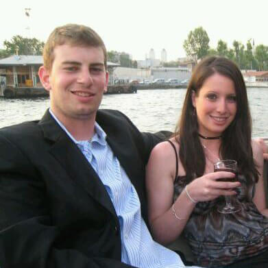 In a marina, a young man wears a suit jacket and no tie puts his arm around a young woman wearing a cocktail dress, holding a wine glass.