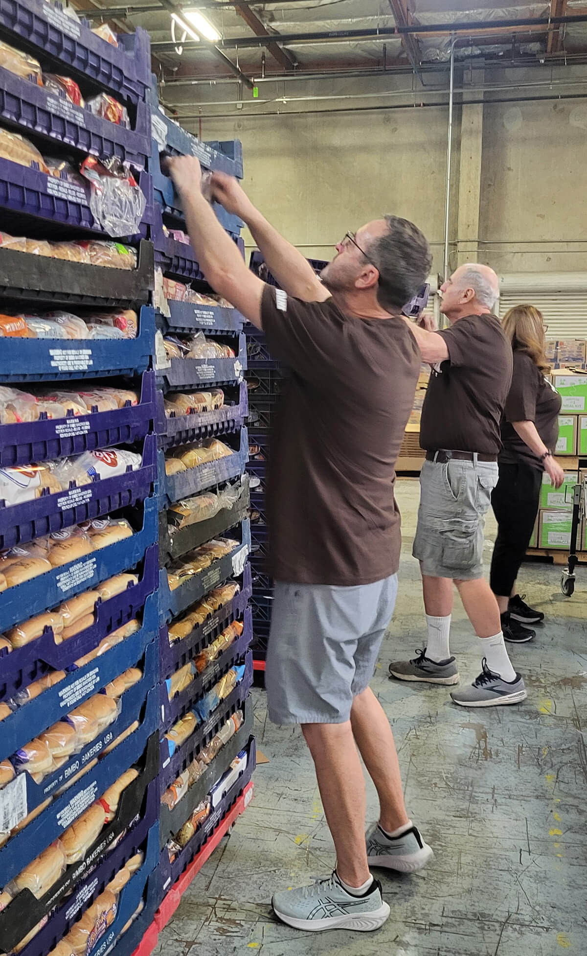 Men in brown tshirts reach up for to blue racks stacked high and filled with bread and rolls.