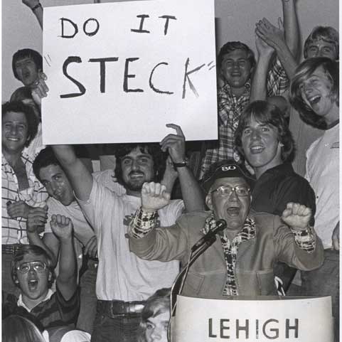 John Steckbeck talks animatedly into a microphone. Amidst the crowd of students behind him, one holds up a, "Do it Steck" sign.