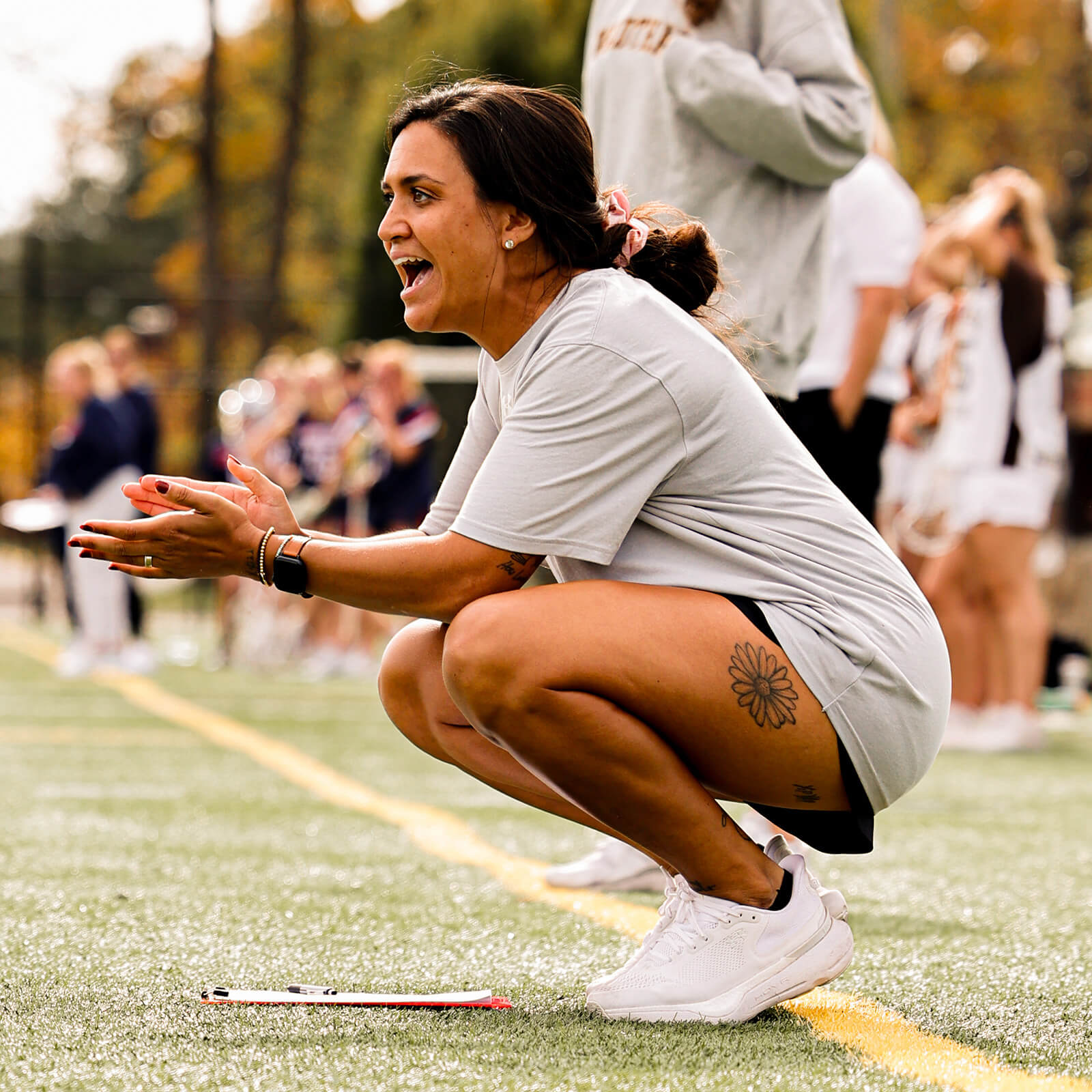 Steph Lazo squats sideline, mouth open, yelling to her team on the field and clapping.