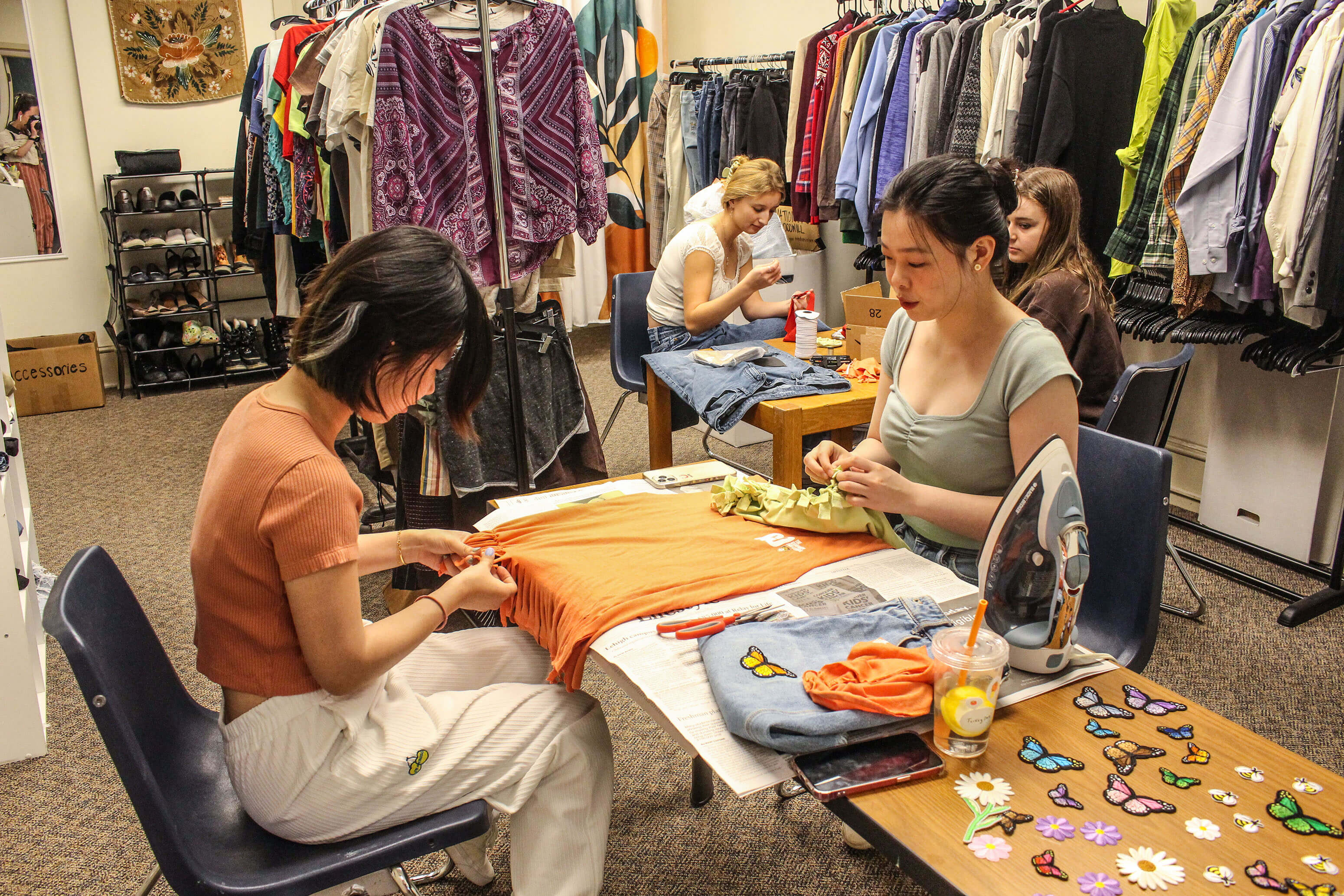 Two students sit at a table to alter thrifted clothes into new, upcycled items by making alterations and adding colorful patches