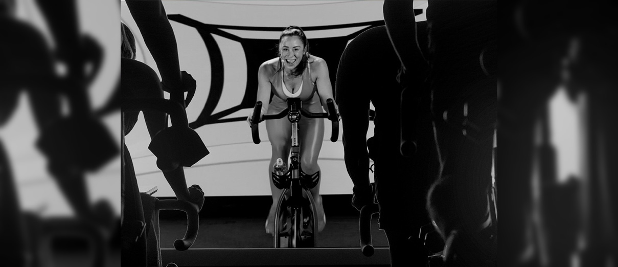 The camera focuses on an enthusiastic spin instructor leads class, represented by shadowed figures on stationary bikes.