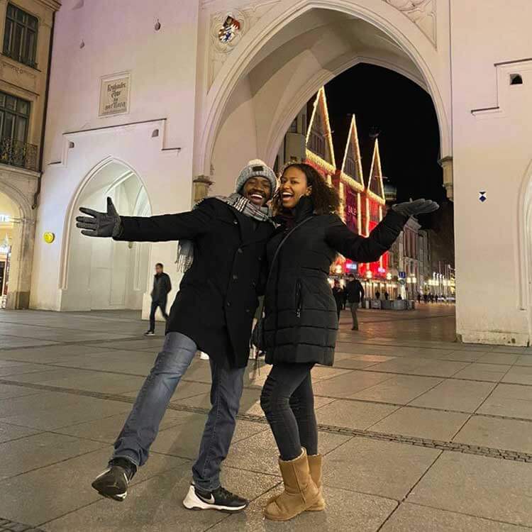 Jones and Taye visit Germany in winter. They wear thick black coats and thrust their arms out by their sides, smiling.