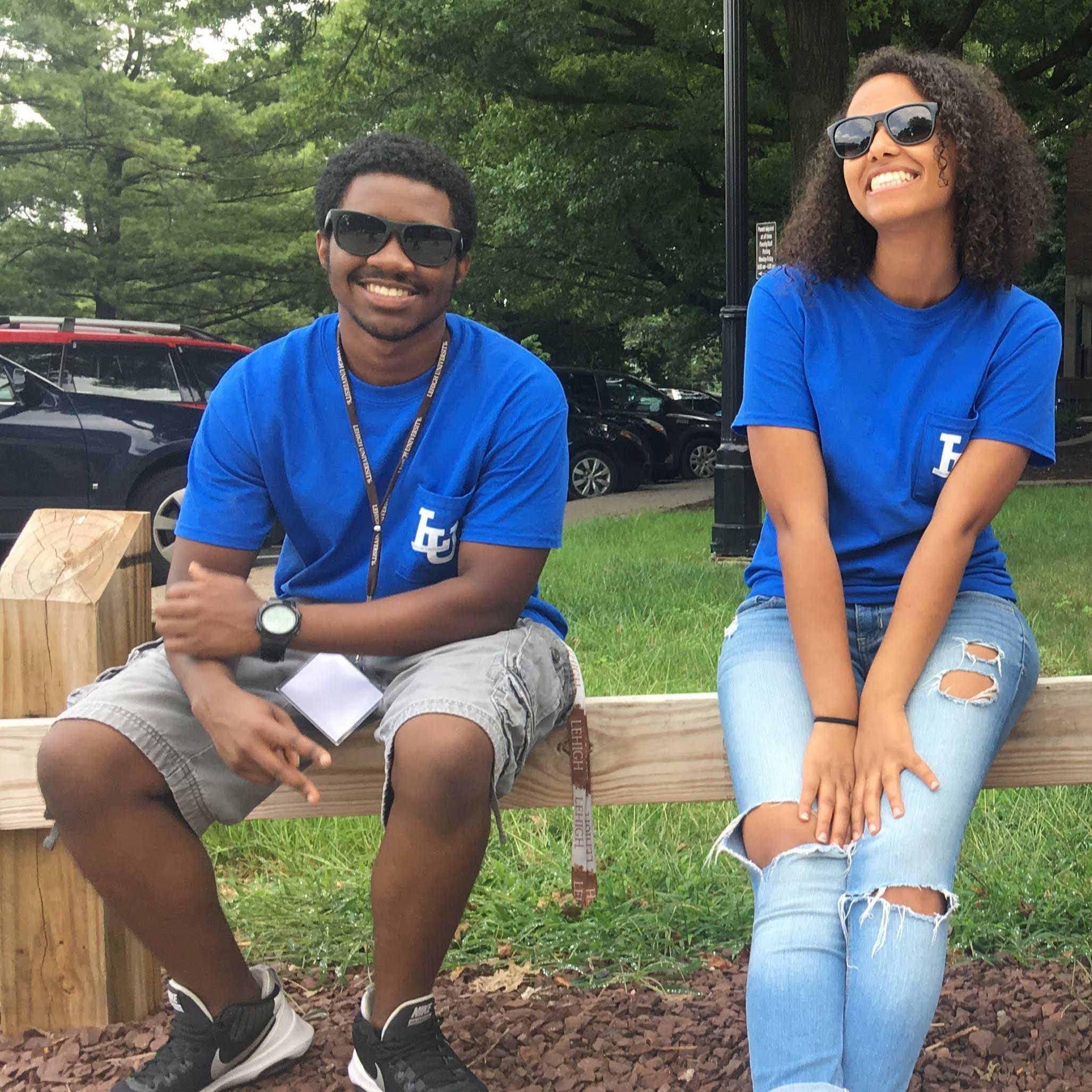 Jones and Taye wear bright blue shirts that mark them as Gryphons and sit outside on a fence. Both wear sunglasses and smile.