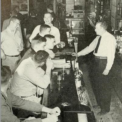 A black and white photo of the inside of the Tally Ho. Several men lean against the bar, laughing and drinking.