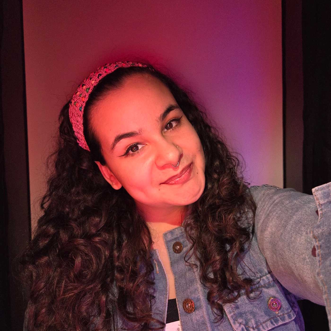 Tanairy wears a bright pink headband, a denim jacket, and striped shirt underneath. The lighting has shades of purple and pink.