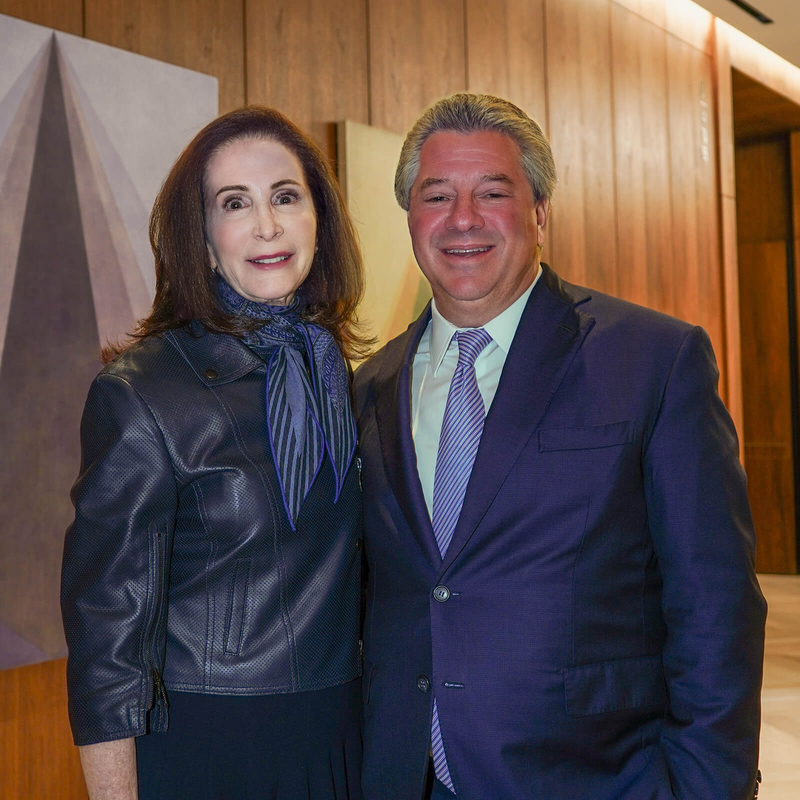 Dressed in corporate attire, Tara Stacom and Marc Holliday stand side-by-side for a photo with wood paneling and geometric artwork behind them.