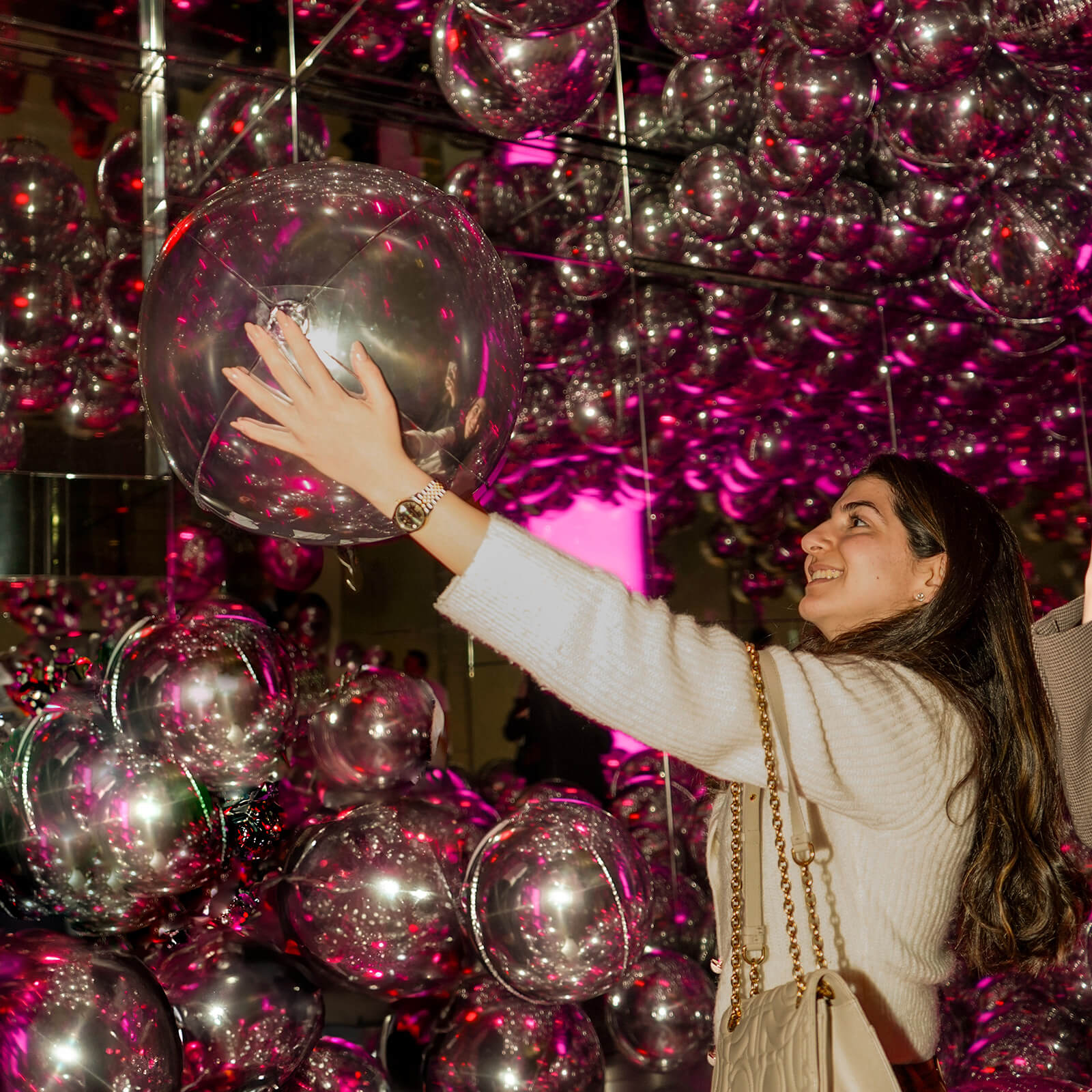 In a dark room filled with balloons illuminated with fuschia lights, a female event attendee reaches up to grab a balloon with a smile.