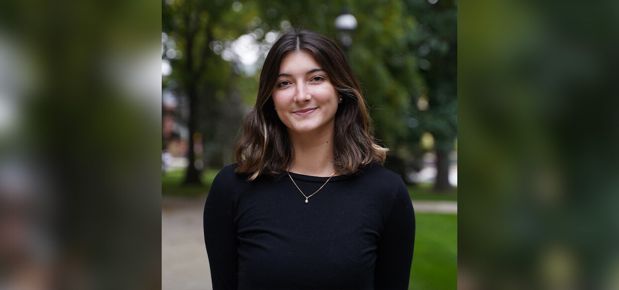 Female student with shoulder length dark hair and a black sweater stands outside on a cloudy day and smiles with a closed mouth for a headshot.