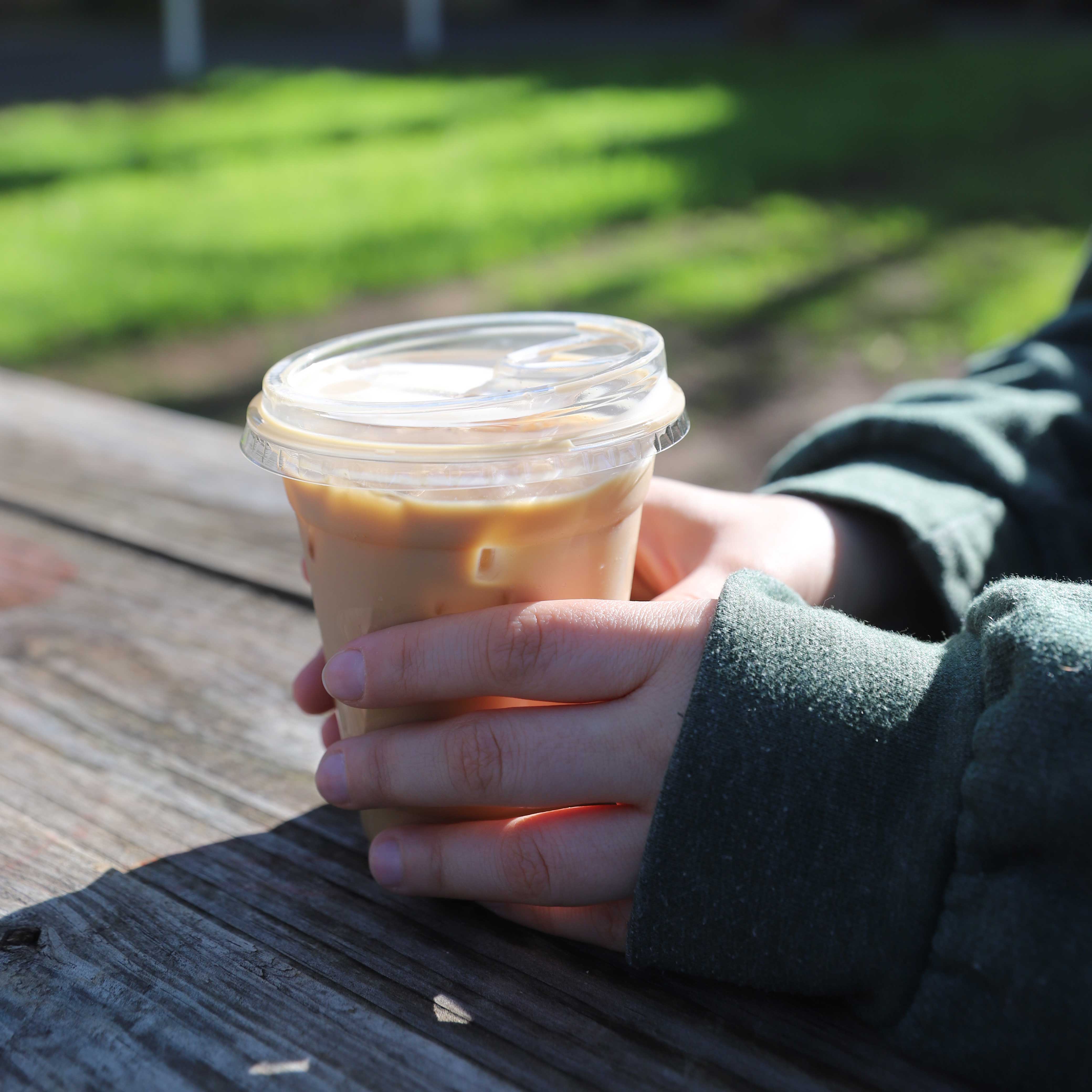 Photo depicts hands holding an iced latte at a picnic bench on a bright, sunny day. 