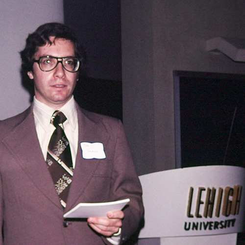 Tom Sulpizio wears a brown suit and holds note cards alongside of a white podium