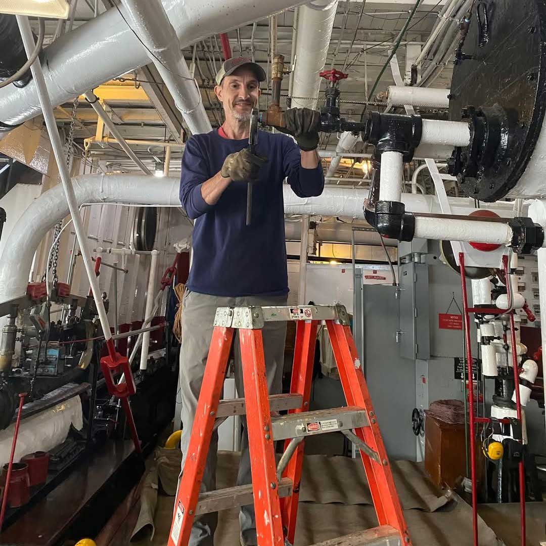 Tom stands on a ladder in the engine room of the Belle and holds a wrench up to a pipe. He wears a dark blue shirt and gloves.