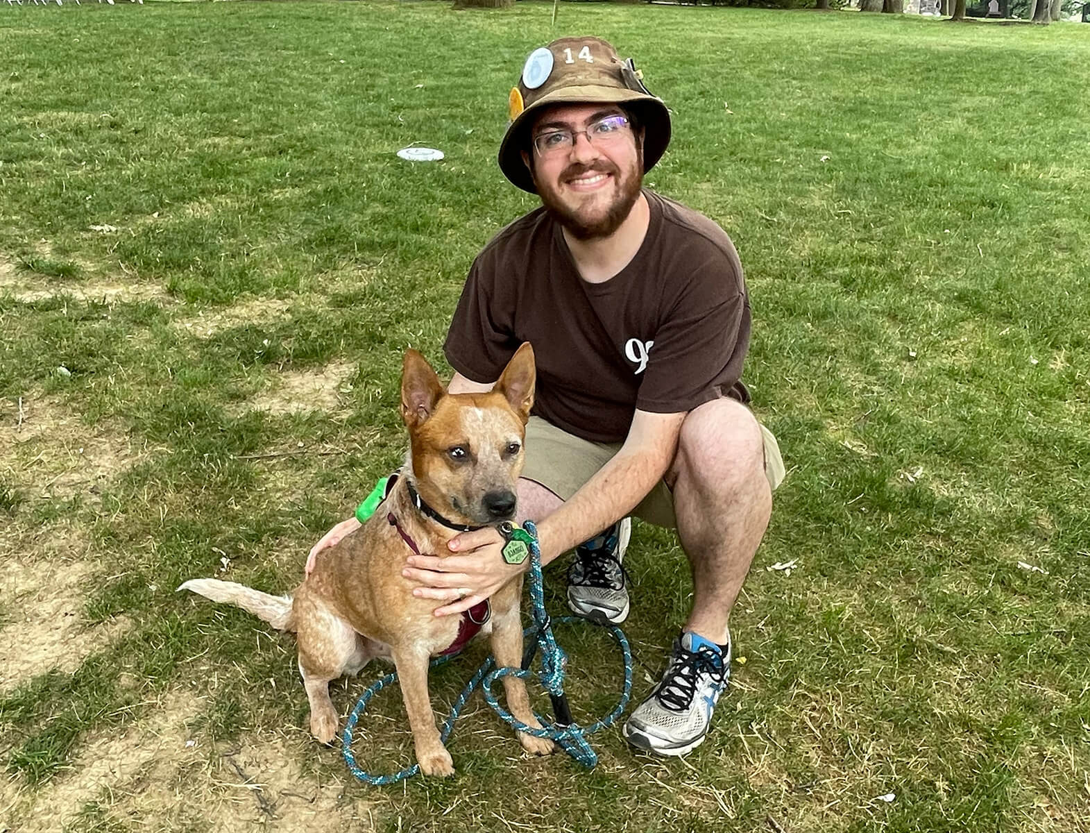 Alumnus wearing a brown t-shirt and bucket hat kneels on the grass beside a sandy colored dog at Reunion 2023.