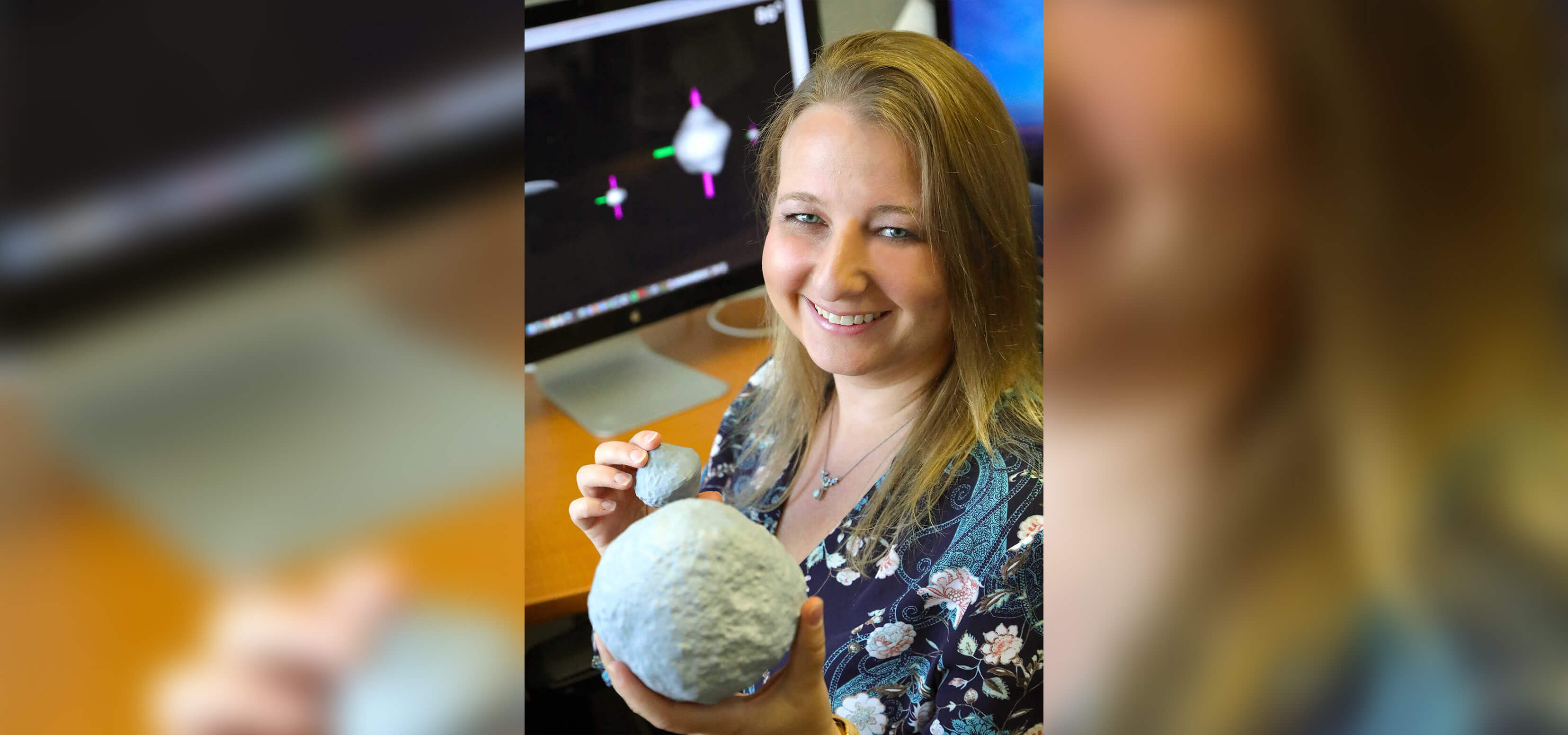 Woman holding two spherical models in front of a computer with a space simulation on screen.