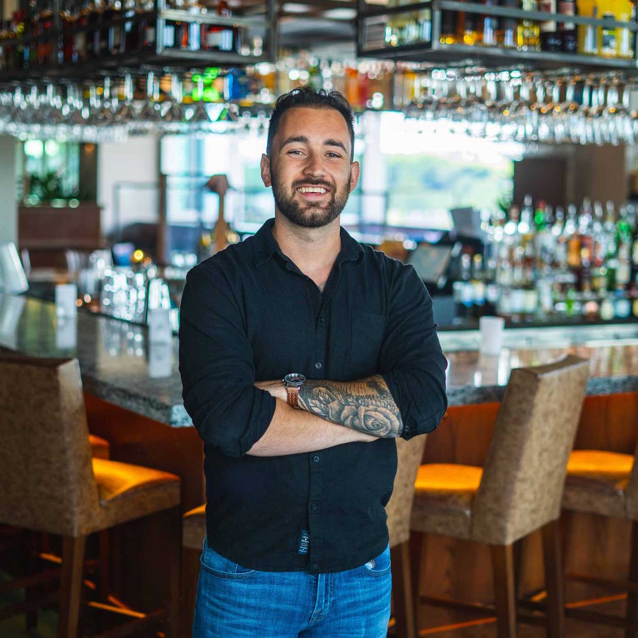 Anthony stands in front of Zest's bar with a smile on his face. His arms are crossed and he wears a black button down with jeans.